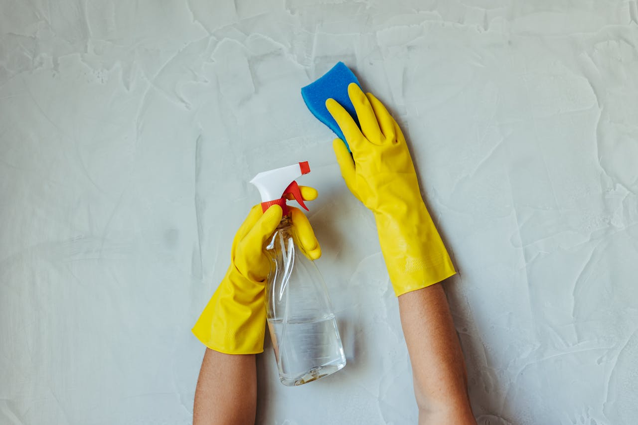 about-img-01 Close-up of hands in yellow gloves cleaning a wall with a spray bottle and sponge.