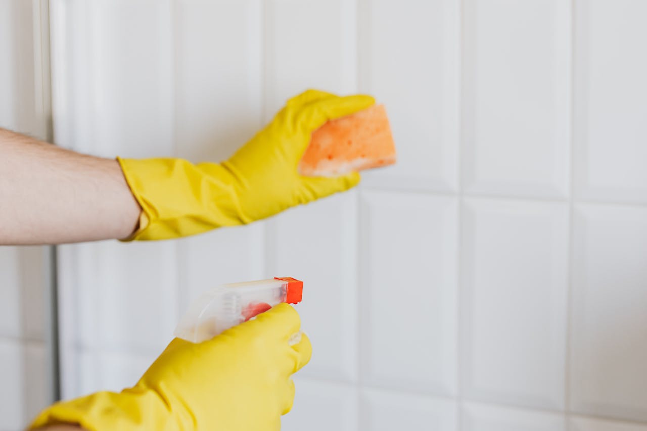 home-img A person wearing yellow gloves cleans bathroom tiles with a sponge and spray bottle.
